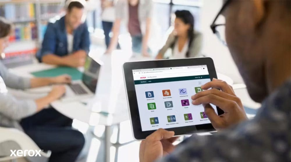 Une personne tient une tablette affichant l'interface Xerox Workflow Central avec diverses icônes d'application, tandis que d'autres personnes travaillent dans l'arrière-plan flou, mettant en évidence l'accès mobile aux solutions de flux de travail.